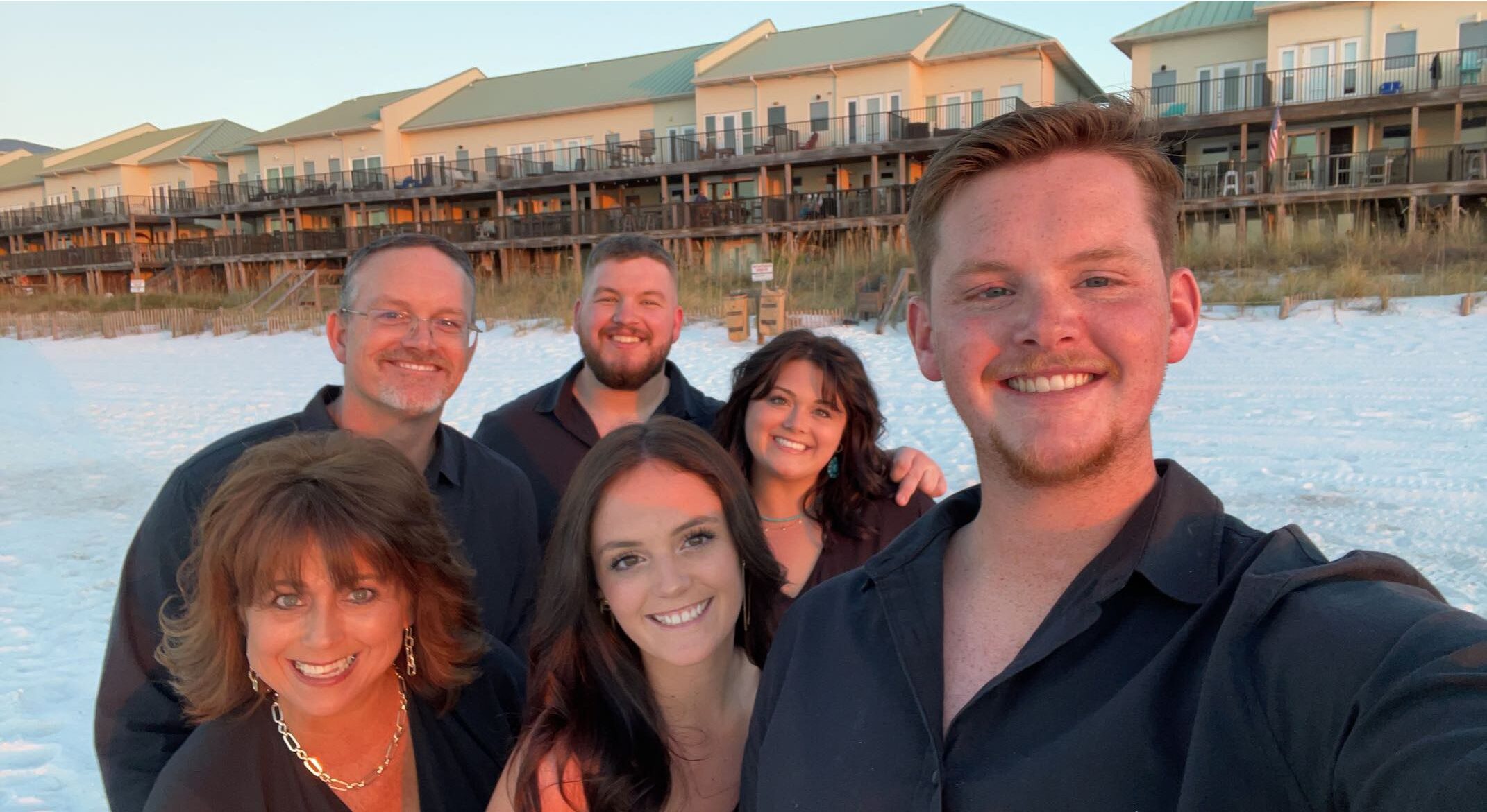 Group selfie of six smiling individuals on a sandy beach at sunset, with beachfront accommodations in the background. Everyone is dressed in black attire, capturing a joyful moment together.