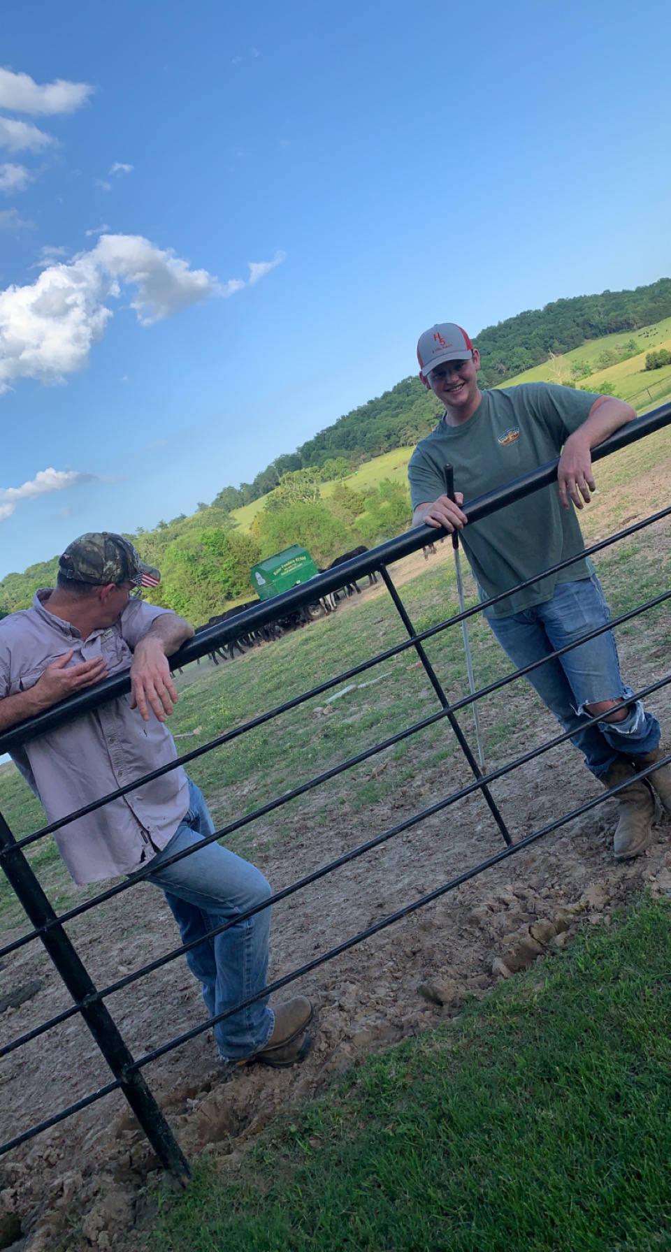 Two men standing by a fence on a farm, with a green truck and cattle in the background. The scene captures a rural landscape with rolling hills and a clear blue sky, highlighting a casual moment in agricultural life.