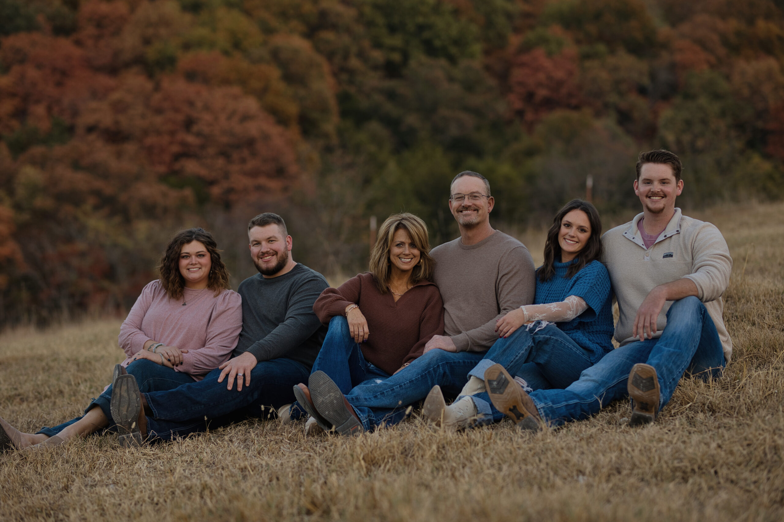 Group of six adults sitting on a grassy field, surrounded by autumn foliage, smiling and enjoying a casual family gathering.
